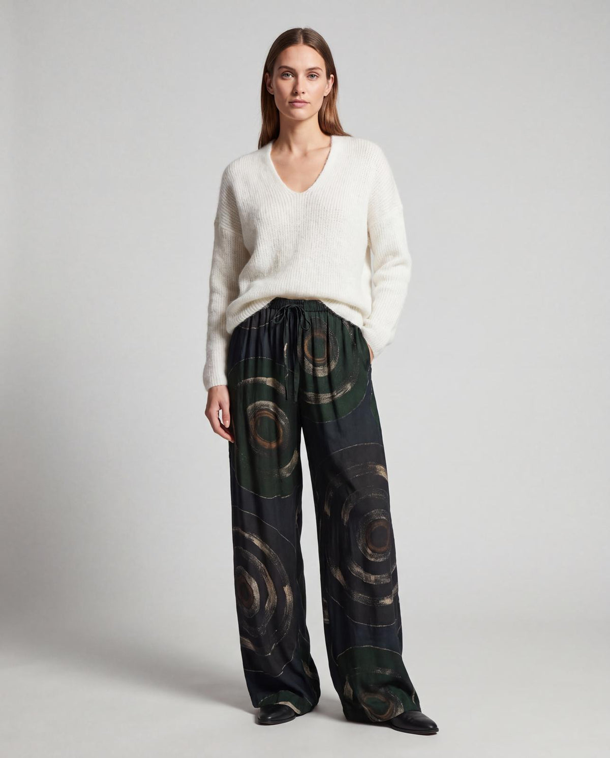Pantalon fluide Cofur en soie de sari upcyclée porté avec un pull col V écru et des bottines noires, grands cercles peints main sur fond noir et vert