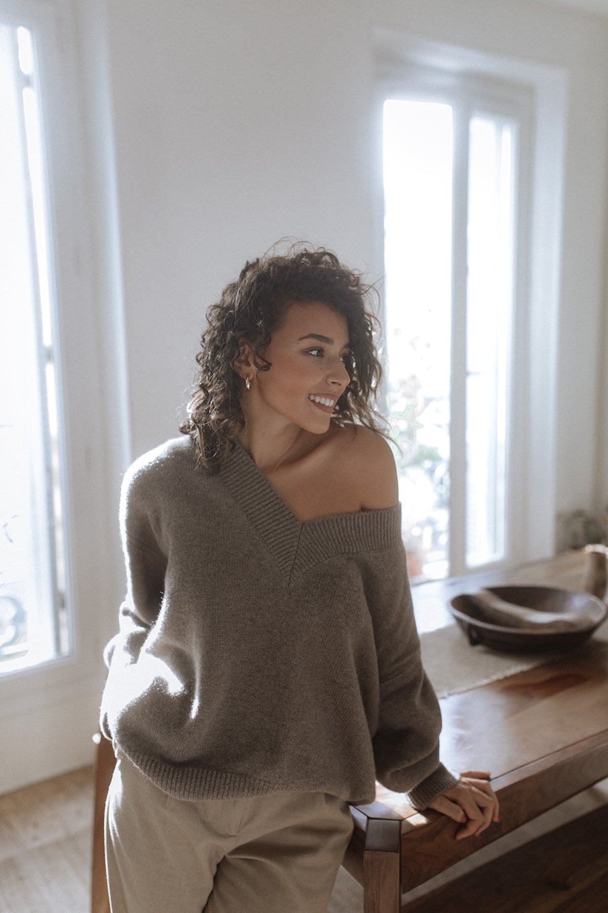 Pull col V Iconic Nuage Cashmere en Capuccino, porté épaule dégagée devant une fenêtre